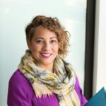 Smiling woman in a purple dress and scarf holding a tablet indoors.
