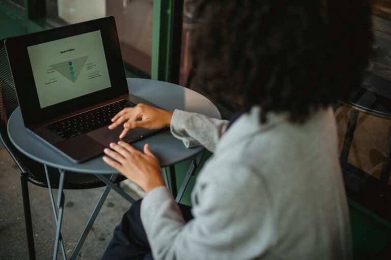 Back view of anonymous ethnic female in suit working with chart on computer while sitting at table in outdoor cafe during remote work