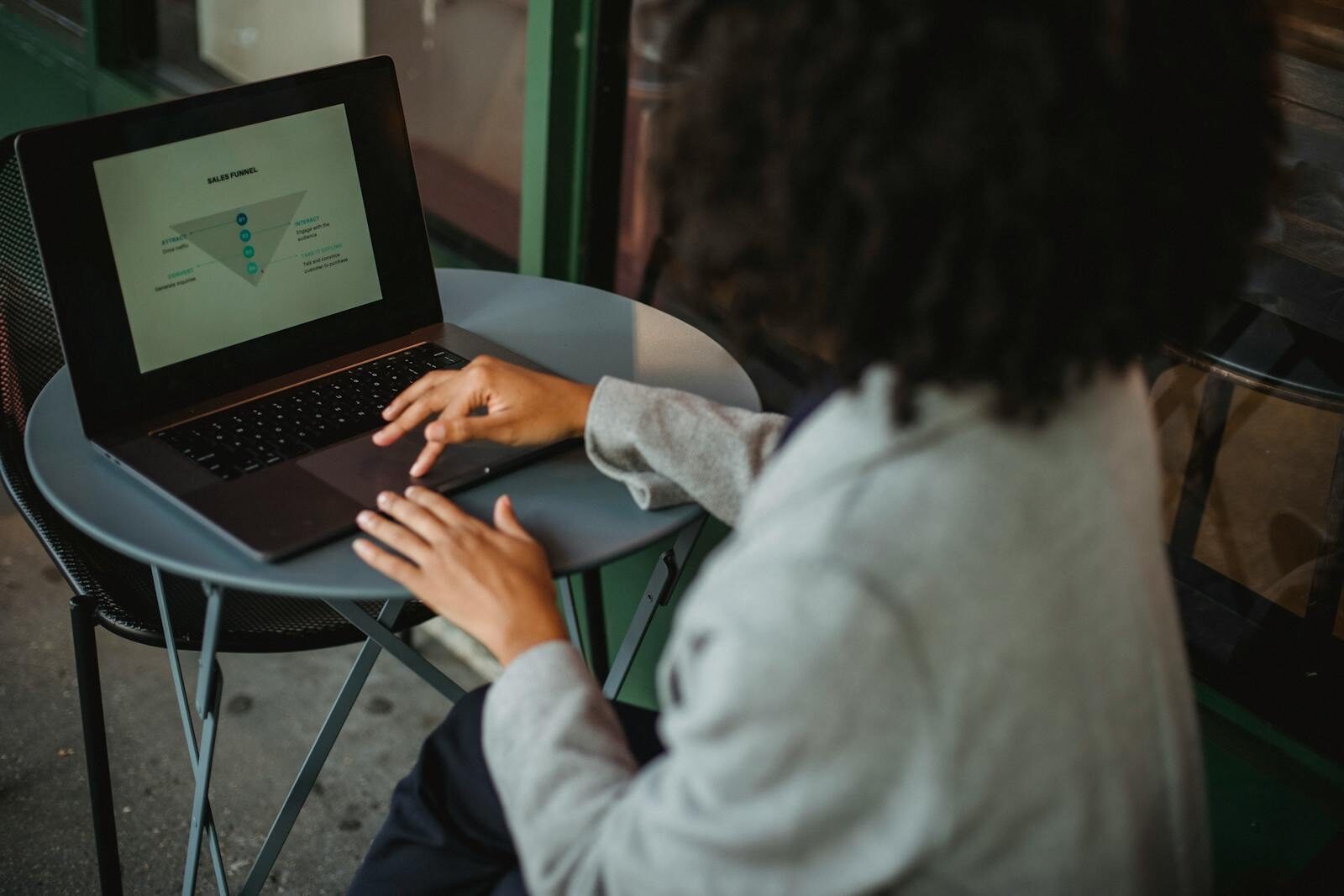 Back view of anonymous ethnic female in suit working with chart on computer while sitting at table in outdoor cafe during remote work