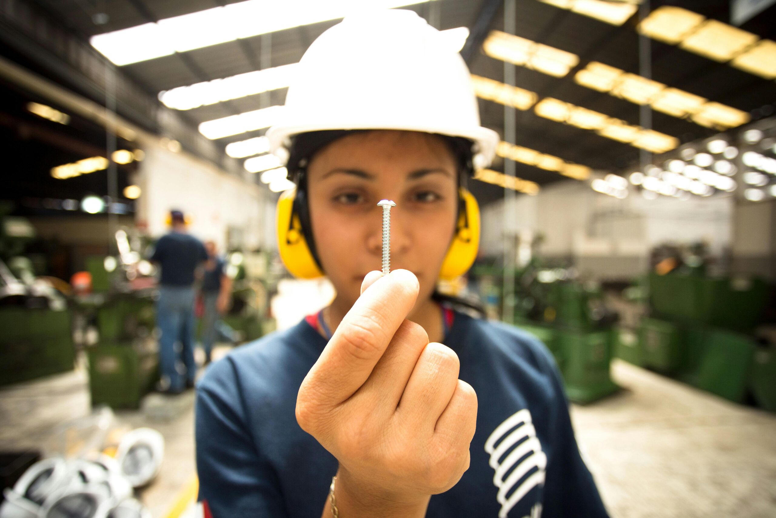 Worker holds a screw in a busy industrial setting, emphasizing precision.