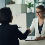 Two businesswomen shaking hands in a modern office setting, symbolizing partnership.