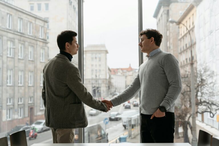 Two businessmen shaking hands in an urban office with city view through large windows.